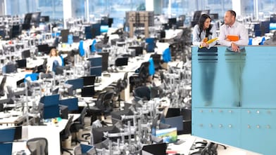 Two people stand on a balcony overlooking a spacious office with desks, computers, and equipment in an open-plan layout.