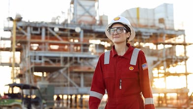 Female Shell worker in red coveralls and safety gear at an industrial construction site, with scaffolding in the background.
