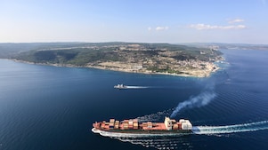 Container ship sailing along a coastal shoreline, leaving a wake across calm blue water.