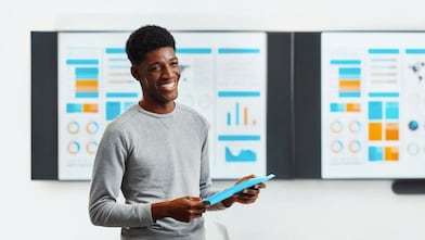 Person holding a blue tablet in front of two large screens displaying colorful charts and graphs, including bar charts and circular diagrams, in a bright office setting.