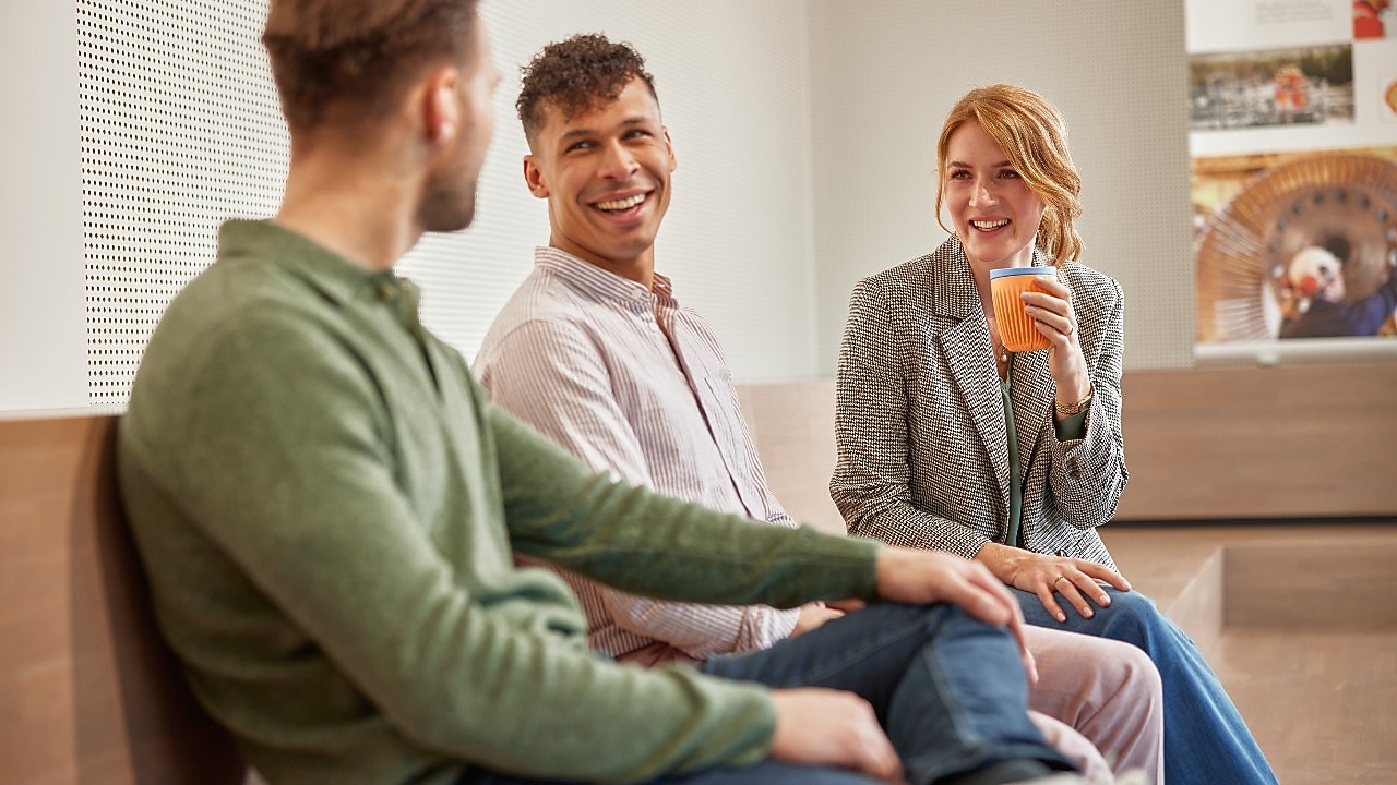Three people sitting on a bench indoors, engaged in conversation; one person holds an orange disposable cup.
