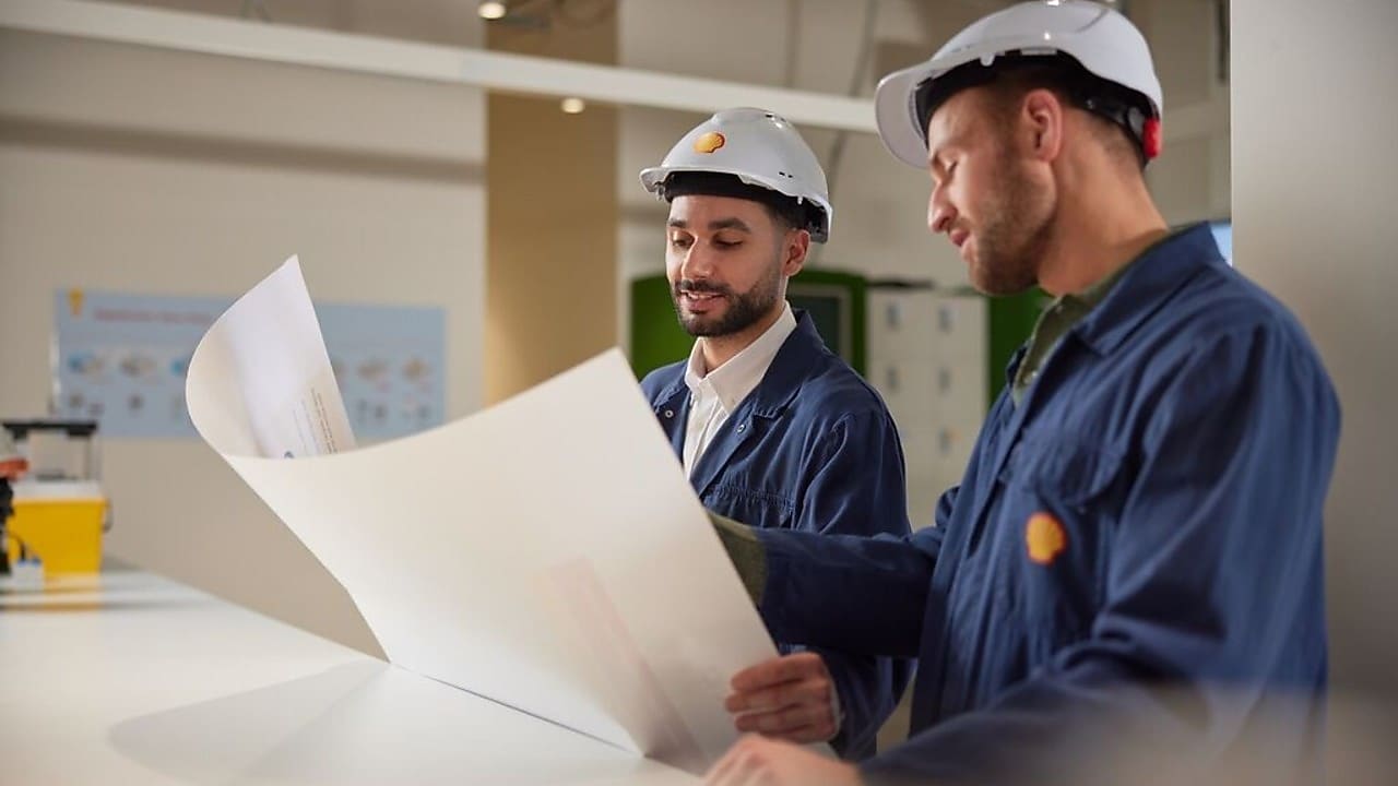 Two individuals wearing white safety helmets and blue work uniforms reviewing large technical drawings in an industrial setting.