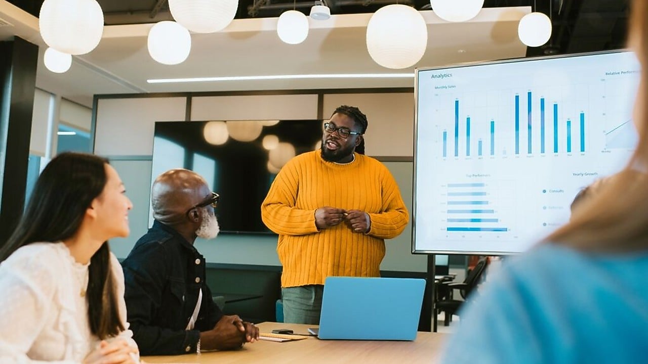 Team gathered in a meeting room with a large screen showing analytics charts. One individual stands near the screen presenting, while others sit around a table with laptops and notebooks.