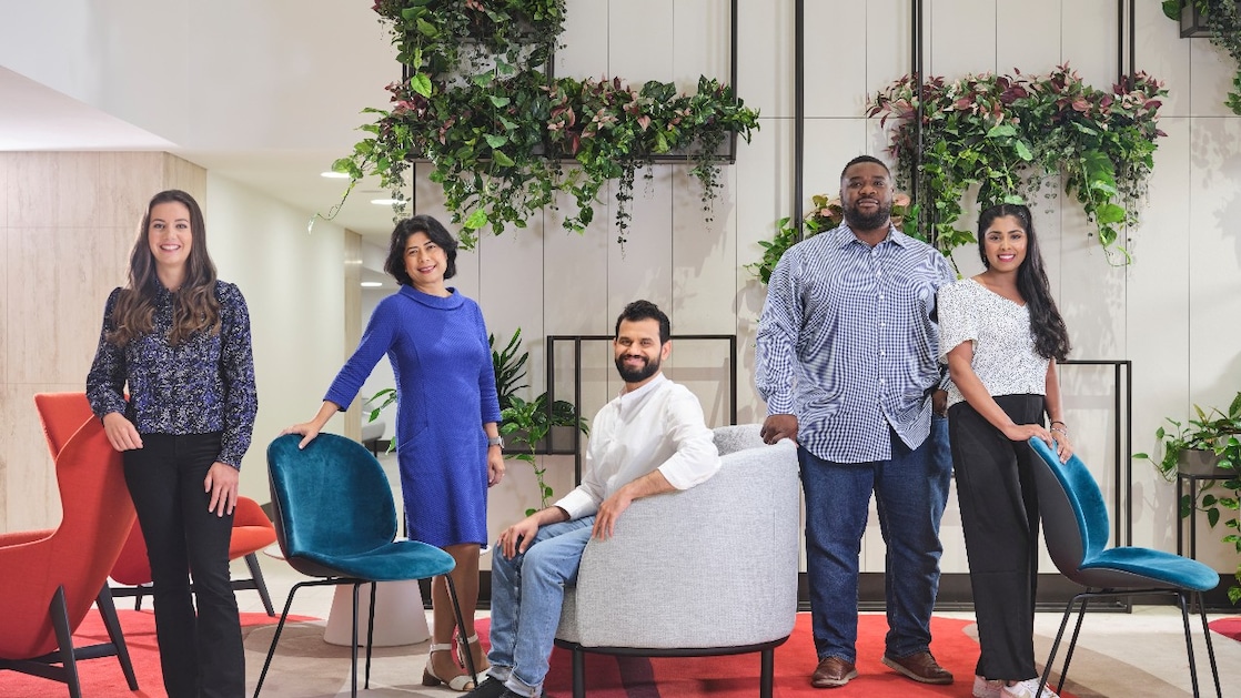 Group of five individuals posing in a modern lounge area with stylish chairs, a red rug, and vertical wall planters filled with greenery.