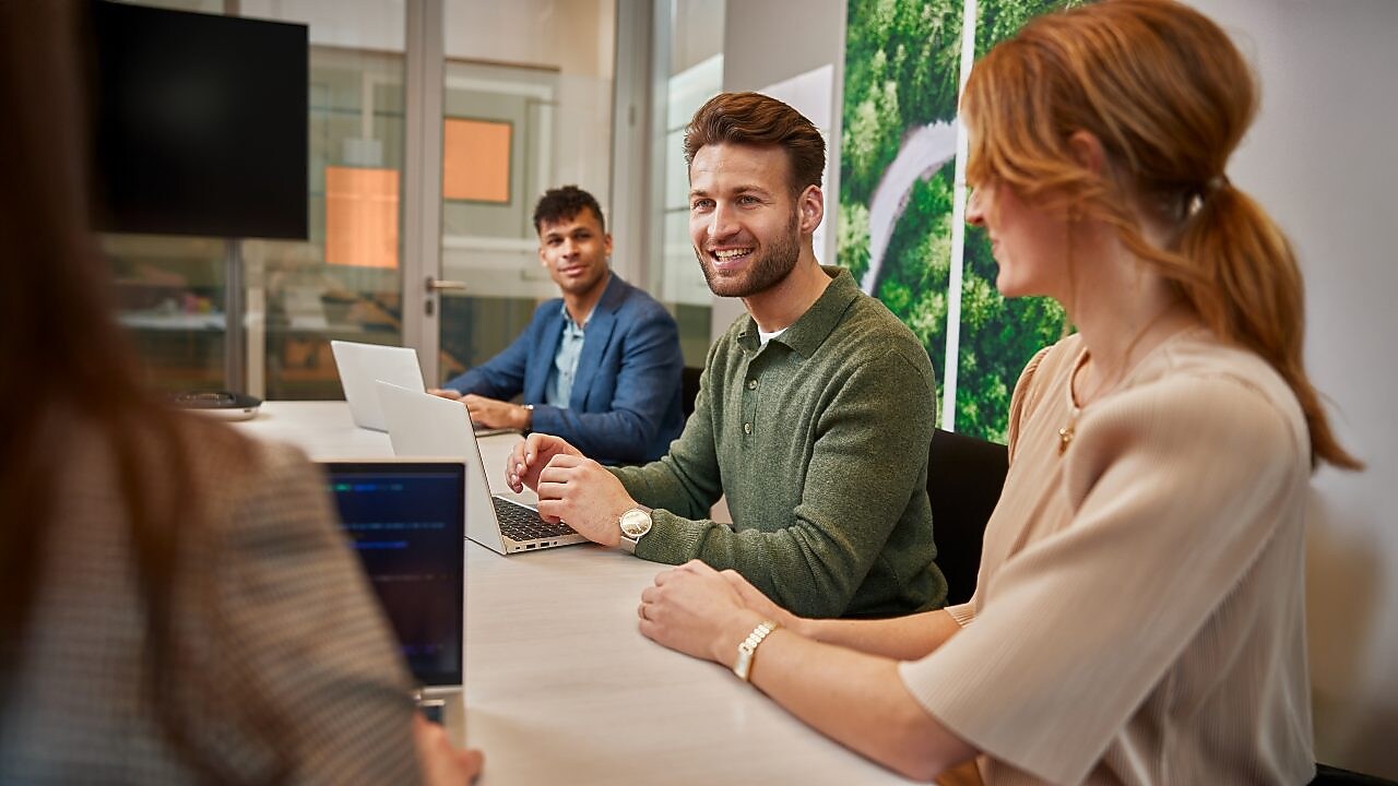 Three people seated around a conference table in a modern office setting, engaged in discussion. Two laptops are open on the table, and a large wall panel with a green aerial view of a forest and winding road is visible in the background