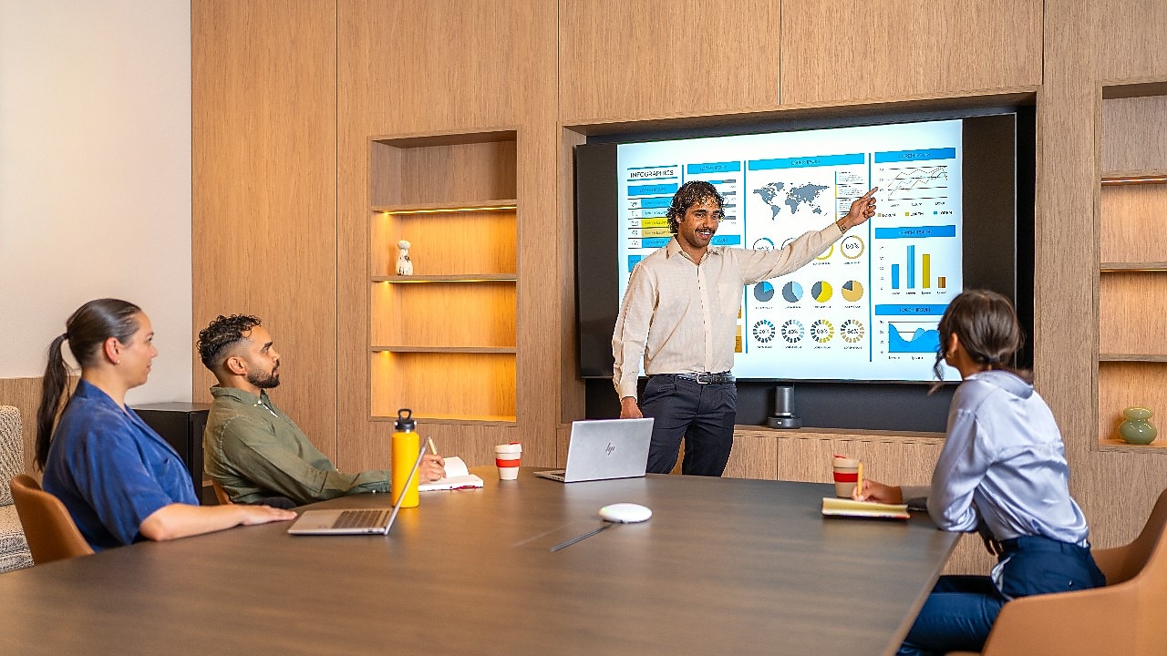 Person standing and pointing at a large screen displaying charts, graphs, and a world map during a presentation in a modern conference room. Three other people are seated at the table with laptops, notebooks, and coffee cups in front of them.