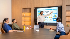 Person standing and pointing at a large screen displaying charts, graphs, and a world map during a presentation in a modern conference room. Three other people are seated at the table with laptops, notebooks, and coffee cups in front of them.