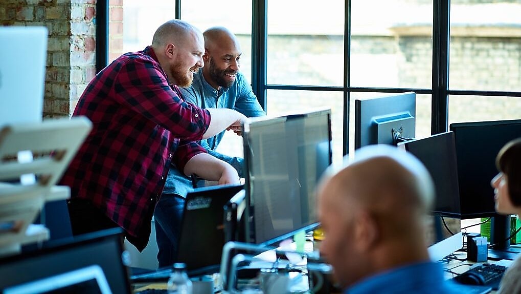 Team members collaborating in a modern office with brick walls and large windows, surrounded by computers and other tech equipment.