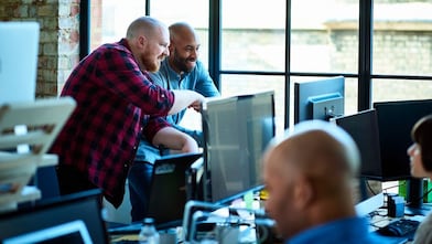 Team members collaborating in a modern office with brick walls and large windows, surrounded by computers and other tech equipment.