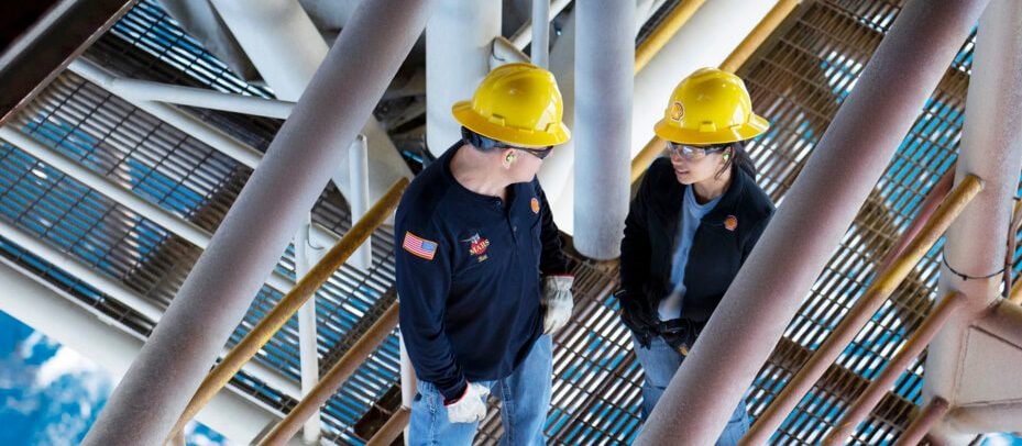 Photograph showing two workers wearing yellow hard hats and safety gear standing on a metal grated platform of an offshore oil rig