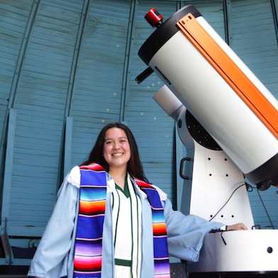 woman in graduation robe in front of telescope 