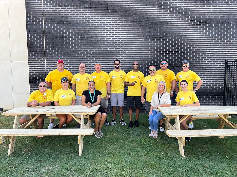 A group of volunteers in a yellow shirt smile in front of newly built picnic benches.