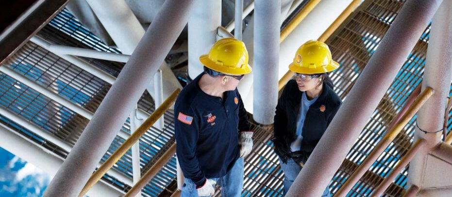 Photograph showing two workers wearing yellow hard hats and safety gear standing on a metal grated platform of an offshore oil rig