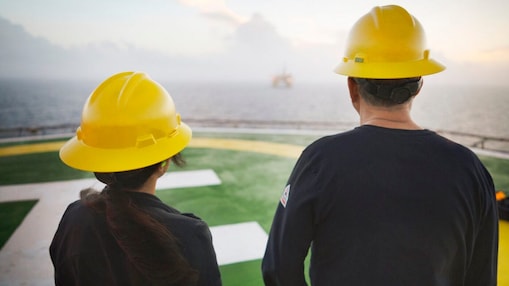 .Two workers wearing yellow hard hats and dark clothing standing on a helipad, facing an offshore oil rig in the distance.
