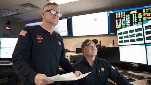 Photograph of two men in a platform control room with multiple computer monitors displaying data and charts.