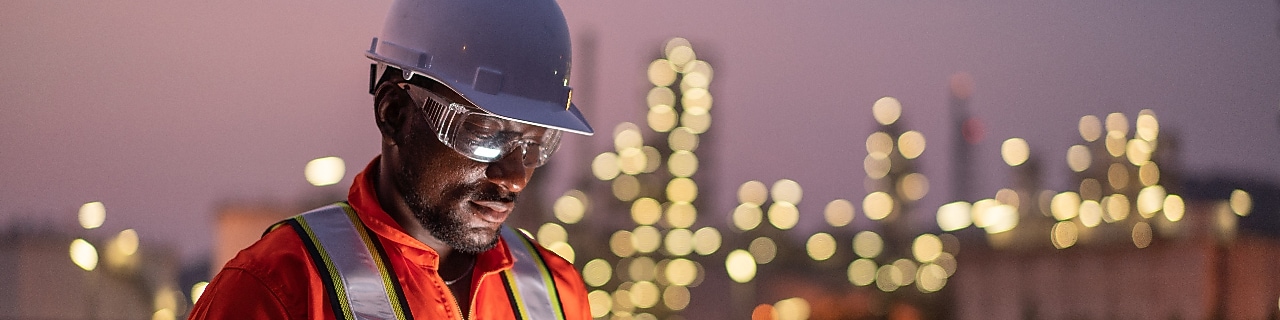 A guy wearing glasses and a helmet looking at a tablet and a refinery in the background