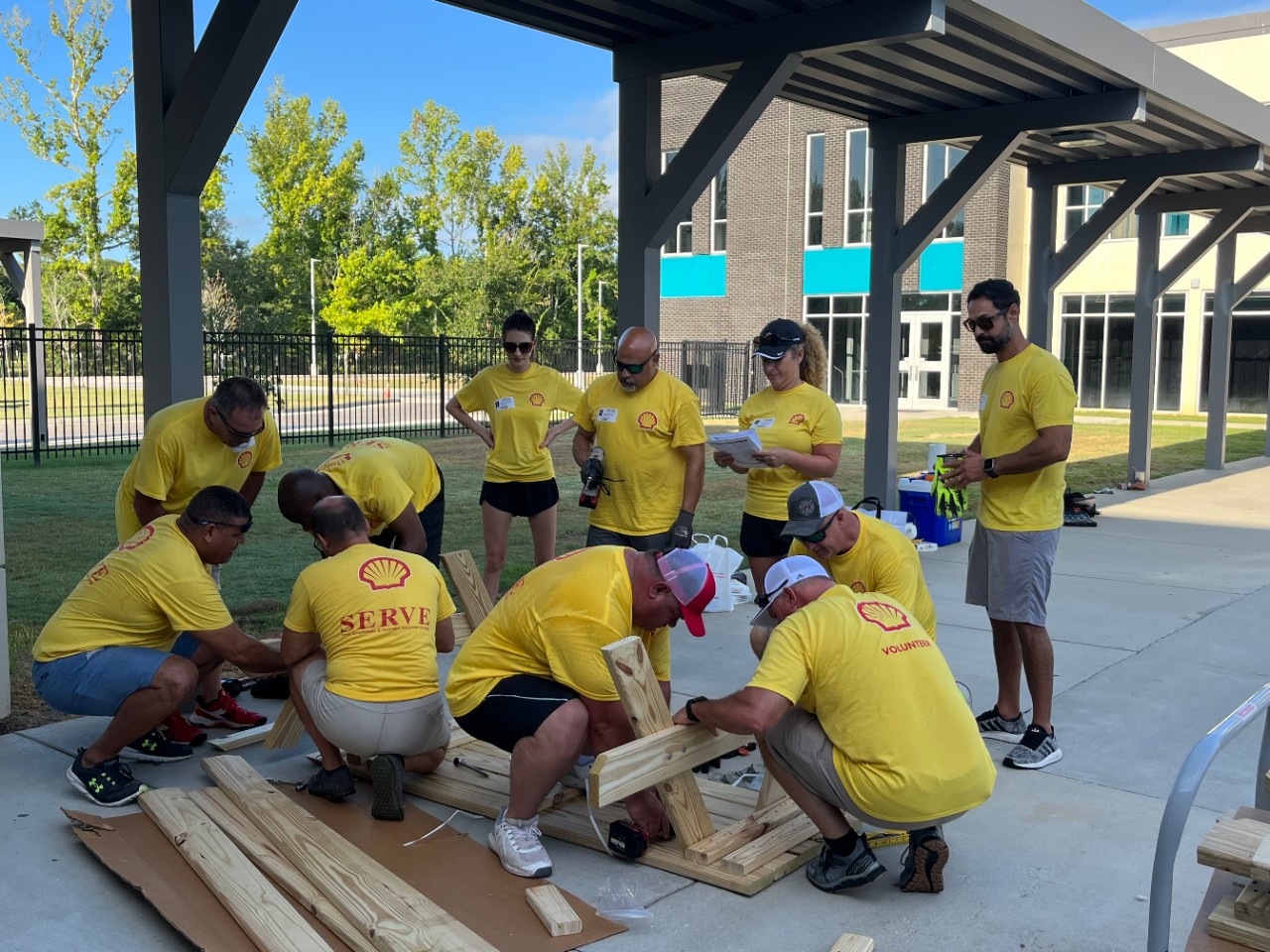 A group of volunteers in a yellow shirt smile in front of newly built picnic benches.