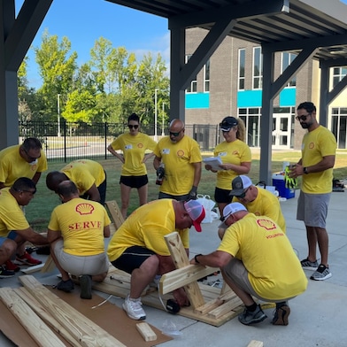 A group of volunteers in a yellow shirt smile in front of newly built picnic benches.