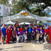 A large group of people pose in front of their company tent for a jambalaya cook-off, a non-profit event.