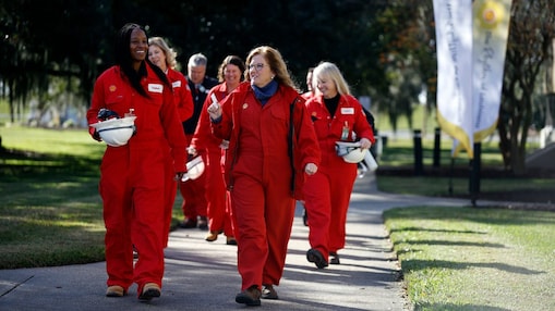 Norco team members, joined by Secretary Giacometto, walk outside at the Norco facility in red coveralls, carrying white hard hats, with Shell flags in the background