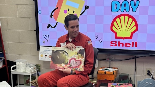 Person in a red Shell uniform sits in a classroom reading a children's picture book aloud. Behind the person, a large screen displays “P.I.E. Reader Day” with the Shell logo and a cartoon book character.