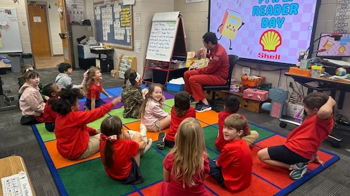 Elementary students sit on a colorful classroom rug while a person in a red Shell uniform reads a picture book at the front of the room. A large display behind the reader shows “P.I.E. Reader Day” alongside the Shell logo and a cartoon book graphic.