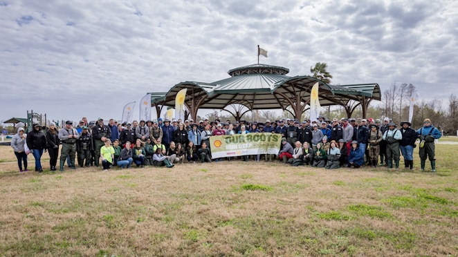 A large group gathers in front of a pavilion holding a banner that reads 'OUR ROOTS RUN DEEP,' surrounded by flags in an open grassy area under a cloudy sky.