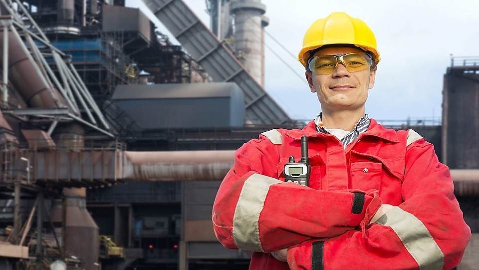 man in red coverall and yellow hard hat standing in front of steel mill
