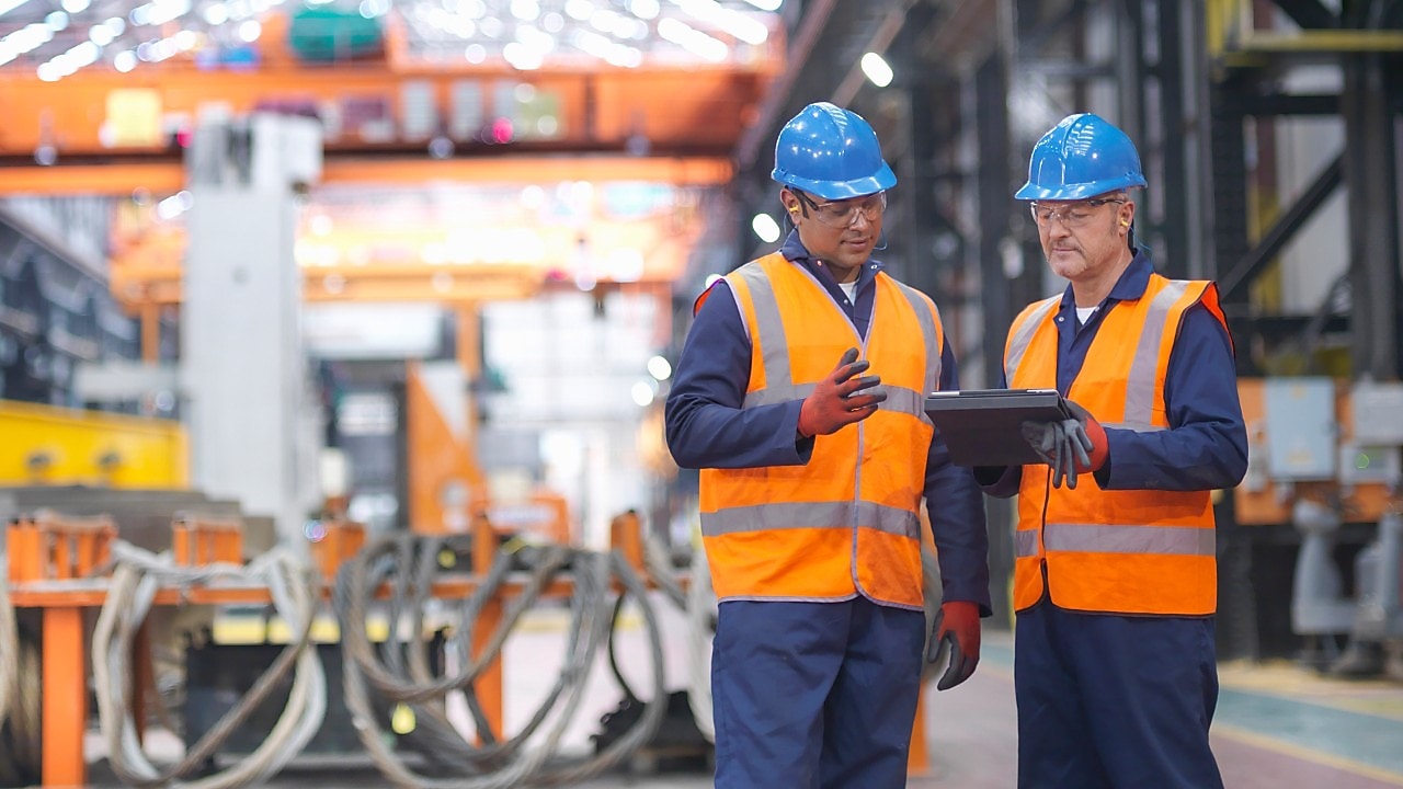 Two industrial workers in hard hats and safety vests reviewing materials at a large manufacturing plant.