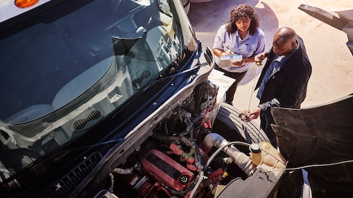 A woman writing notes standing next to a man checking the engine oil on a heavy-duty truck.