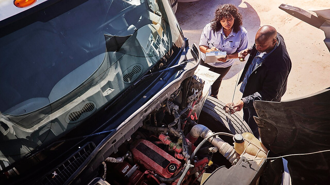A woman writing notes standing next to a man checking the engine oil on a heavy-duty truck.