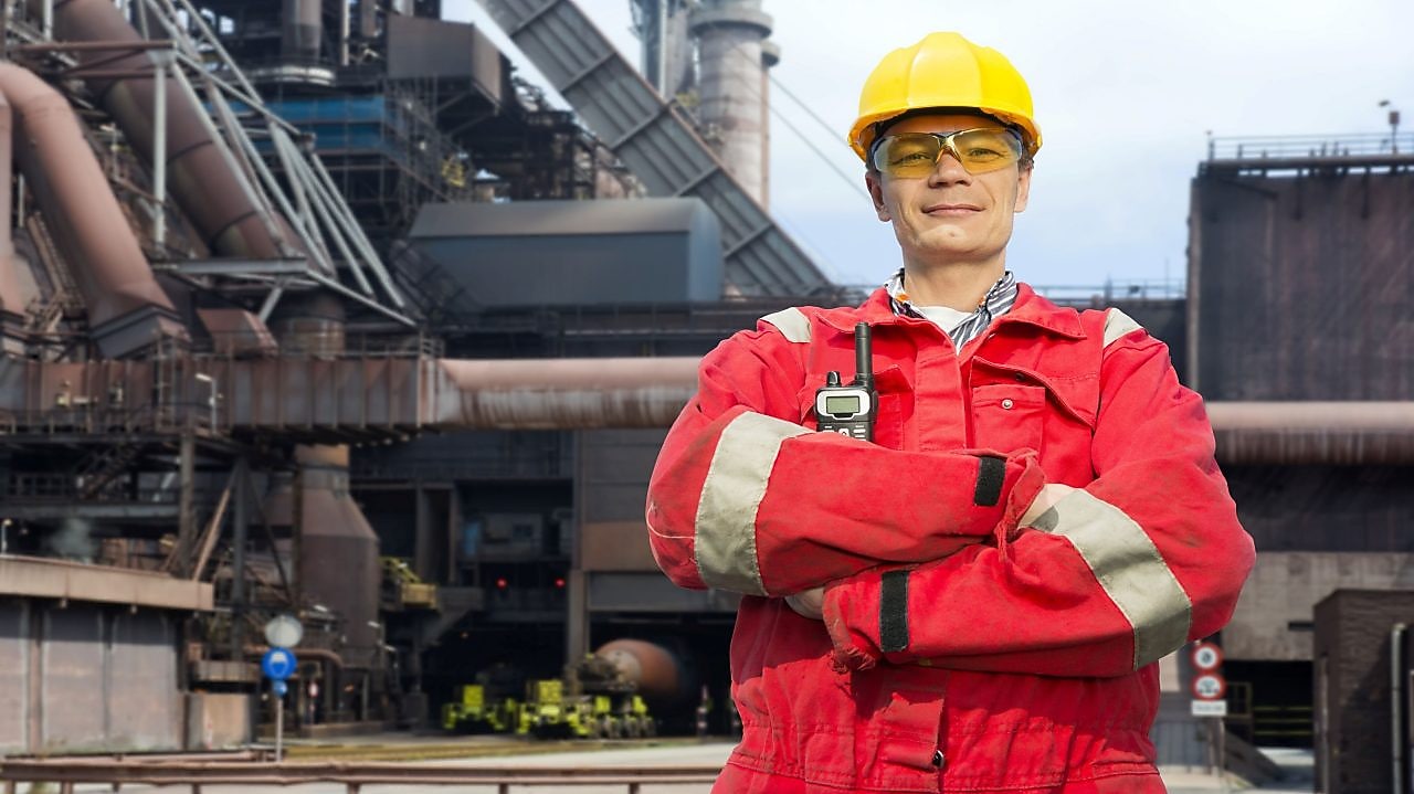 man in red coverall and yellow hard hat standing in front of steel mill
