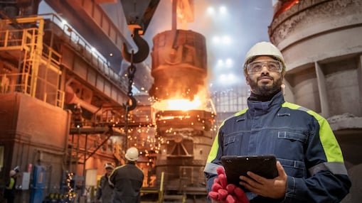 a man in PPE with iPad and steel making process in the background