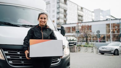 Portrait of smiling female worker carrying box while standing against delivery van in city