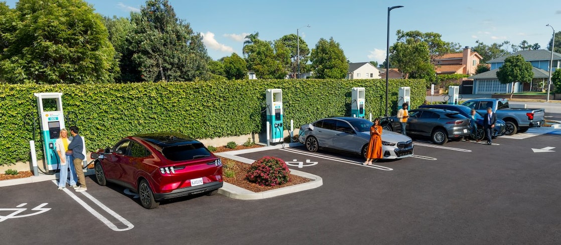 Multiple cars charging at a Shell Recharge hub while drivers move about the site.